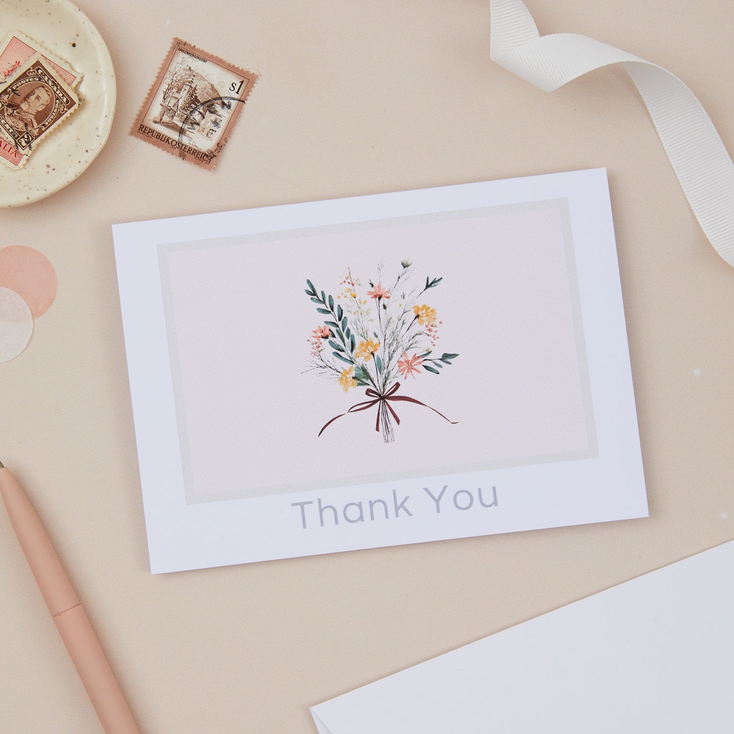 A thank you card with a floral bouquet illustration, placed on a desk with a pen, ribbon, and other desk accessories.