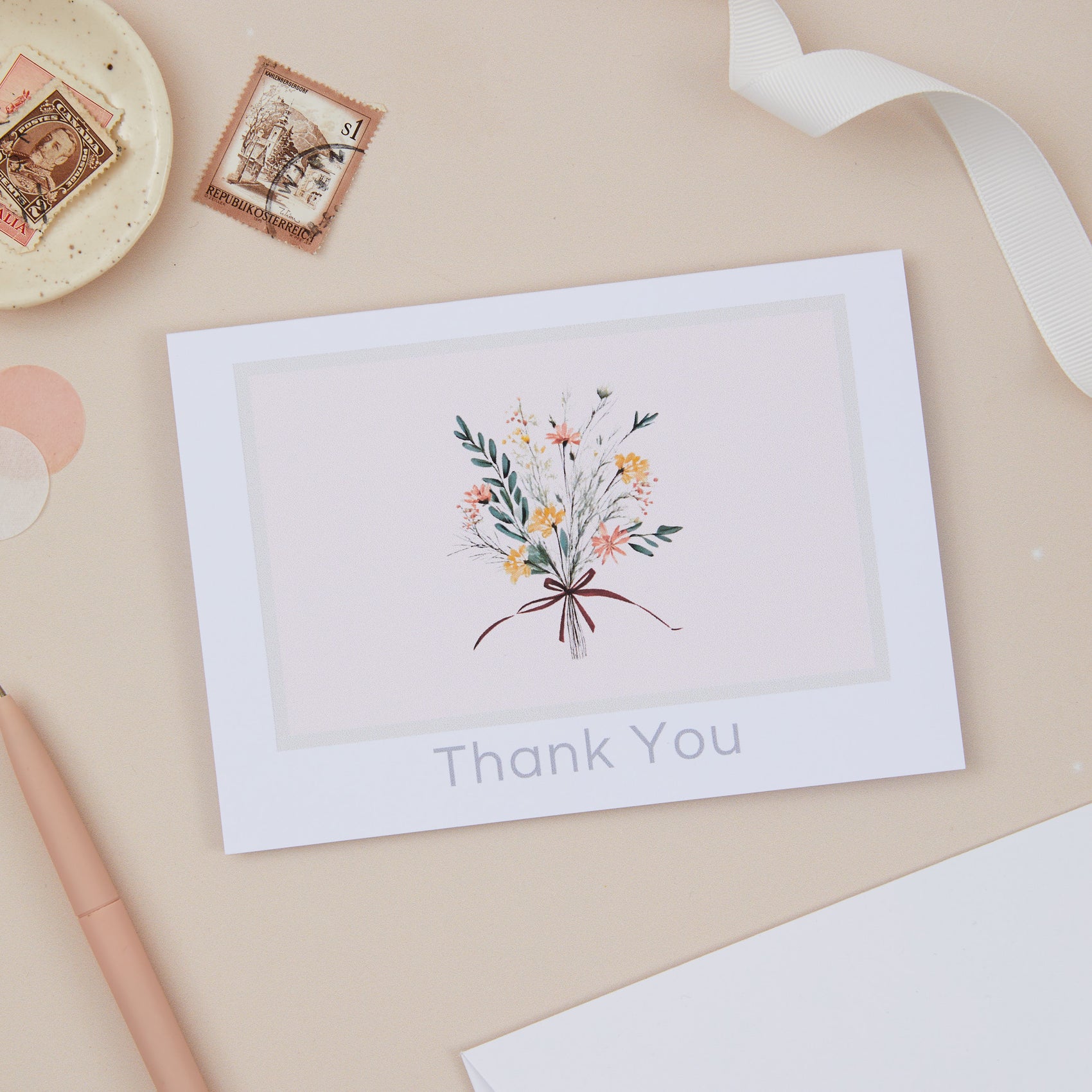 A thank you card with a floral bouquet illustration, placed on a desk with a pen, ribbon, and other desk accessories.