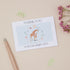 Floral Giraffe Baby Gift Thank You Card Set. A thank you card with a baby giraffe design, surrounded by floral elements, placed on a neutral background with a pen and decorative flowers around it.