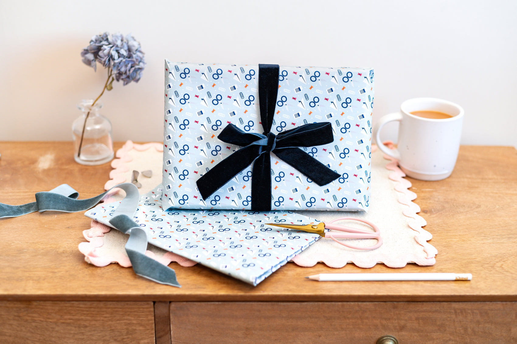 Gift Box wrapped in light blue wrapping paper with scissors & stationery pattern on a wooden table.