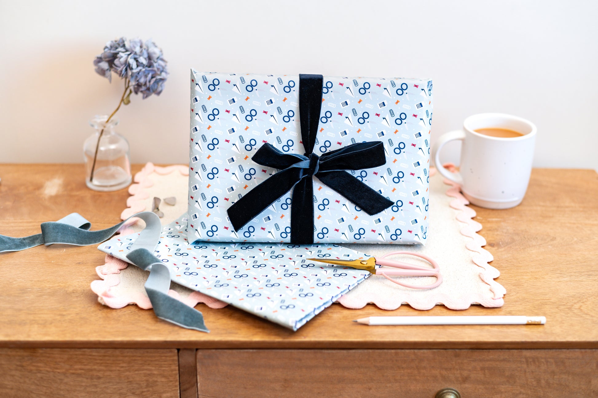 Gift Box wrapped in light blue wrapping paper with scissors & stationery pattern on a wooden table.