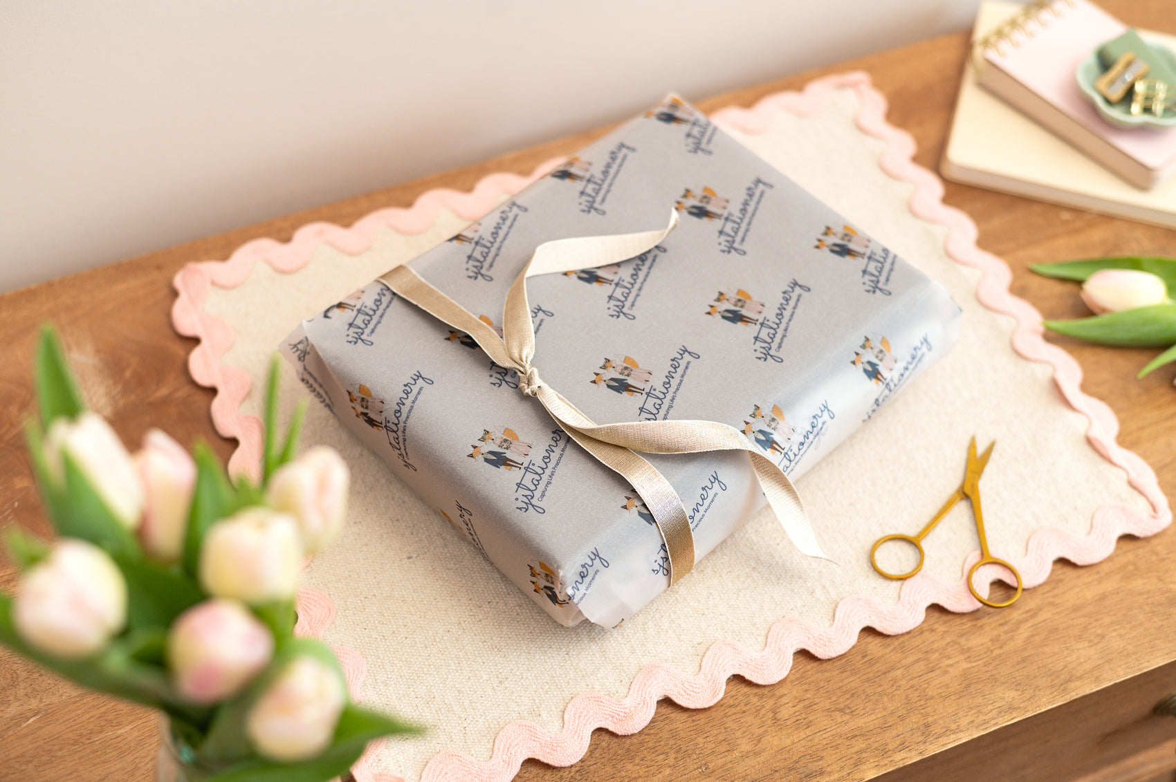 Gift wrapped in branded Sjstationery Paper with a gold ribbon on a wooden surface with flowers and scissors.