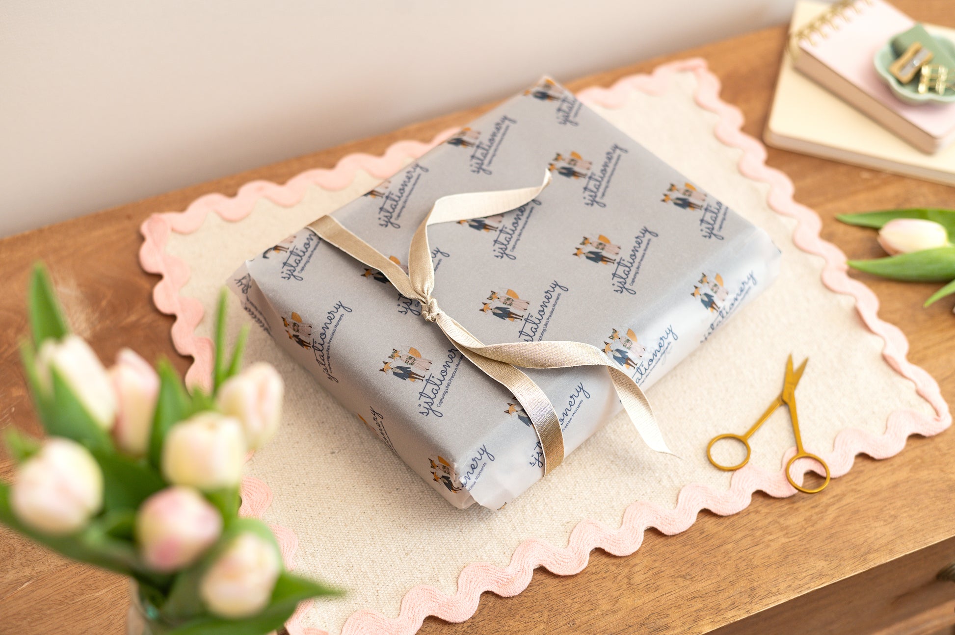 Gift wrapped in branded Sjstationery Paper with a gold ribbon on a wooden surface with flowers and scissors.