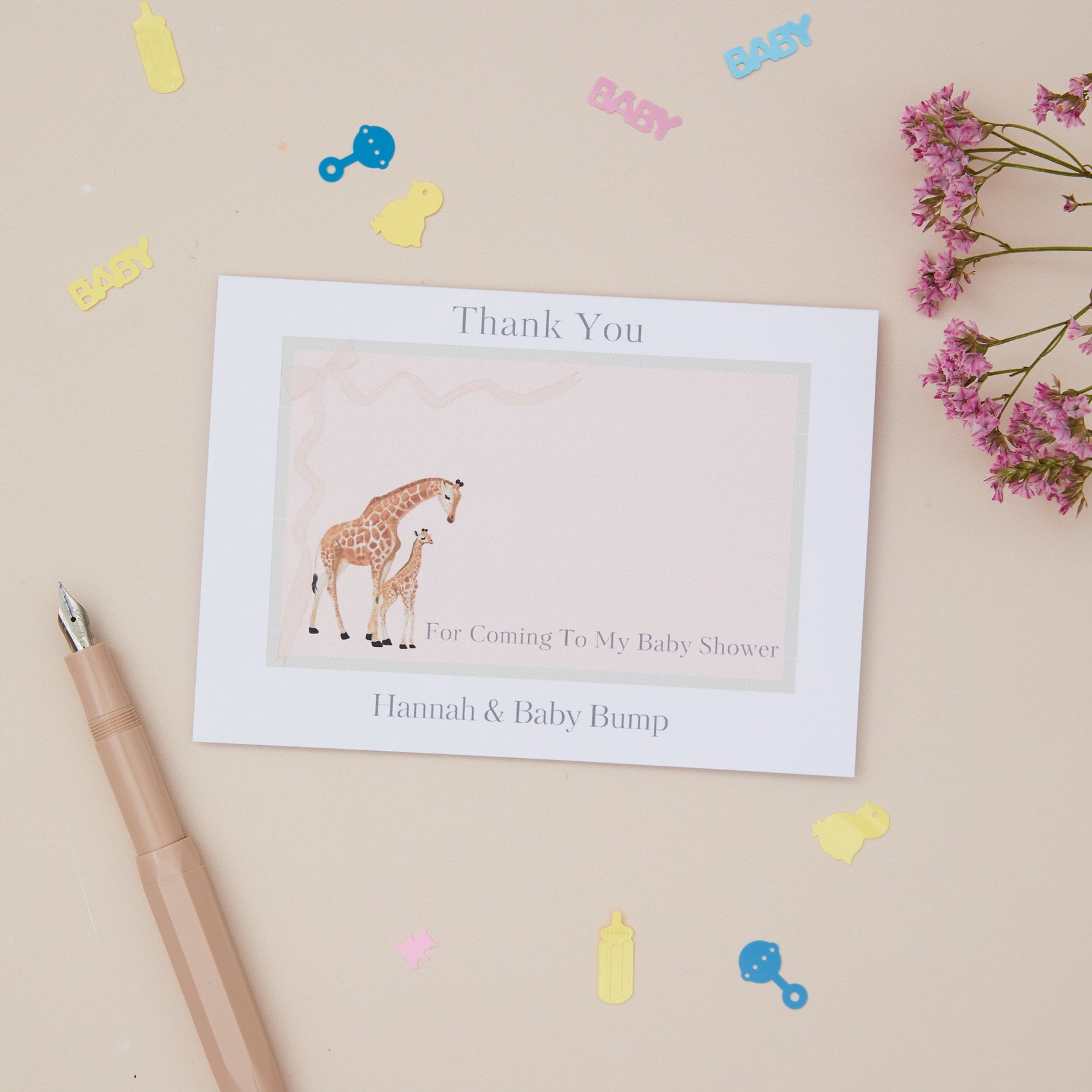 A personalised baby shower thank you card with a cute giraffe and baby design, customised with a name, placed next to a pen, on a background with decorative elements.