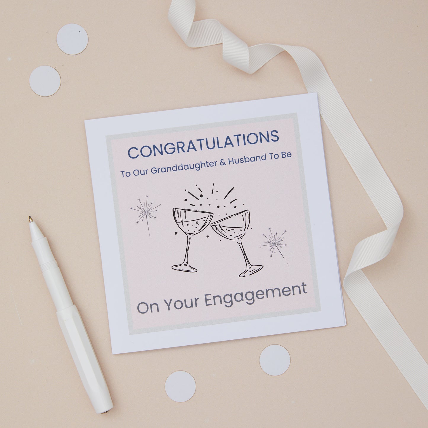 A Granddaughter  engagement congratulations card with two glasses toast design, including two celebratory glasses, accompanied by sparklers. The card is presented on a neutral background, with a decorative ribbon, confetti & pen. The Card features the text " Congratulations to our granddaughter & husband to be - on your engagement.