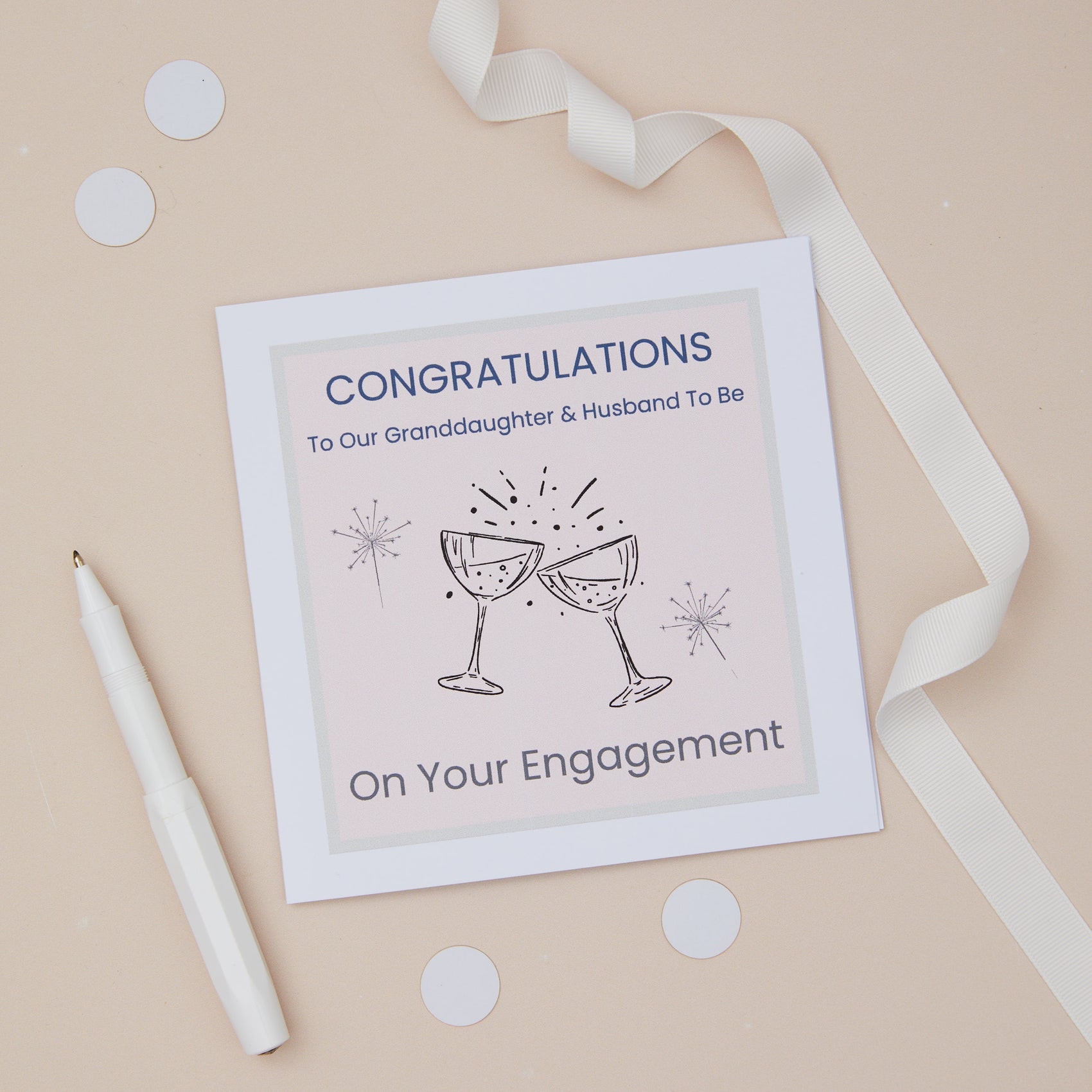 A Granddaughter  engagement congratulations card with two glasses toast design, including two celebratory glasses, accompanied by sparklers. The card is presented on a neutral background, with a decorative ribbon, confetti & pen. The Card features the text " Congratulations to our granddaughter & husband to be - on your engagement.