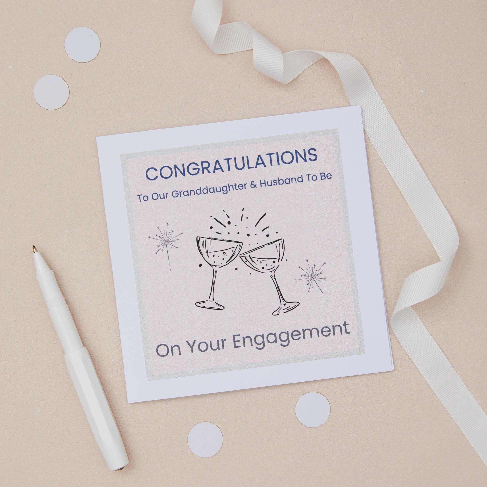 A Granddaughter  engagement congratulations card with two glasses toast design, including two celebratory glasses, accompanied by sparklers. The card is presented on a neutral background, with a decorative ribbon, confetti & pen. The Card features the text " Congratulations to our granddaughter & husband to be - on your engagement.