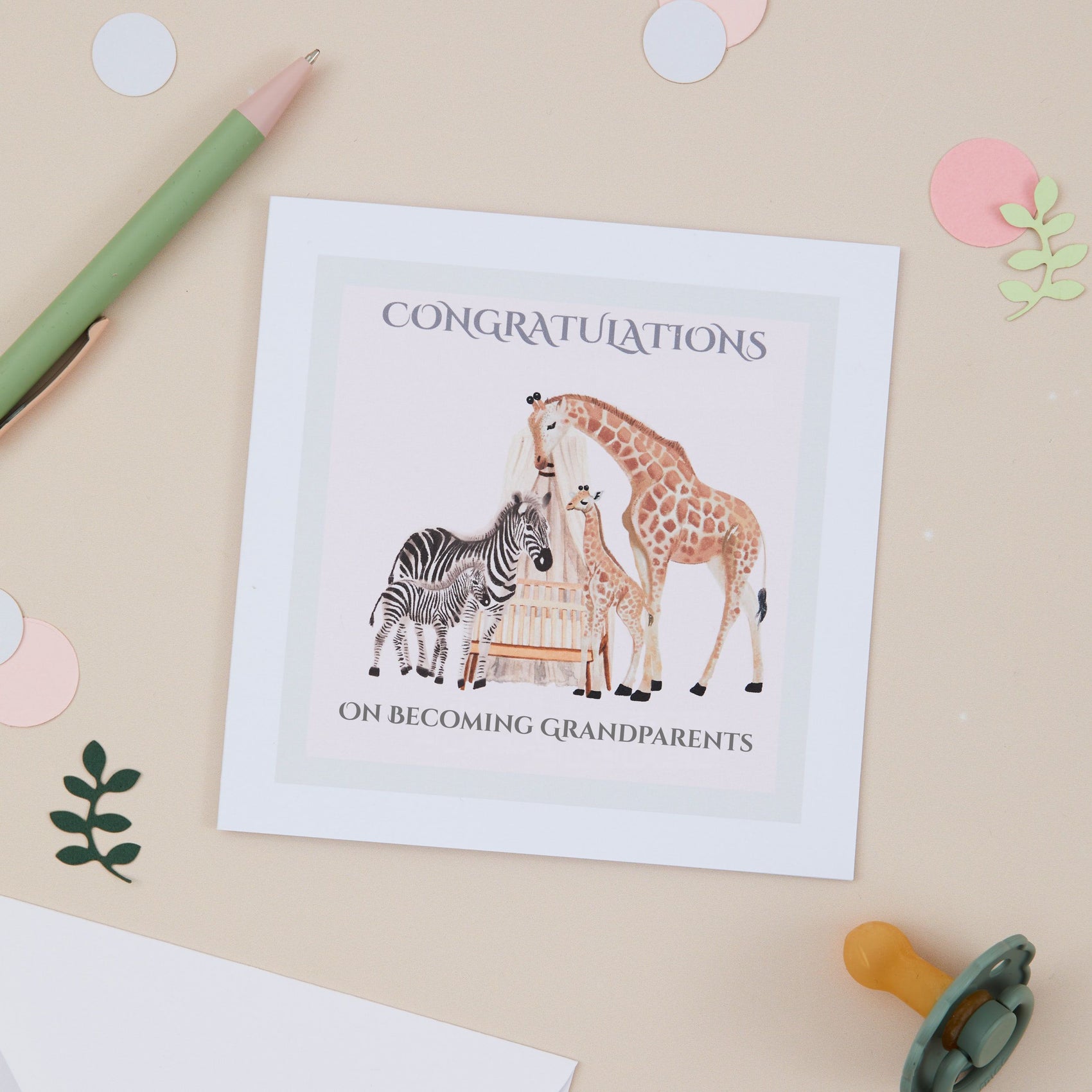 Greeting card with Zebra, Giraffe & their young surrounding a Cot with a pale pink background. The Square card is against a Beige background and there is a pen and a Babies Dummy in the background. The card has the text 'Congratulations on becoming grandparents'.