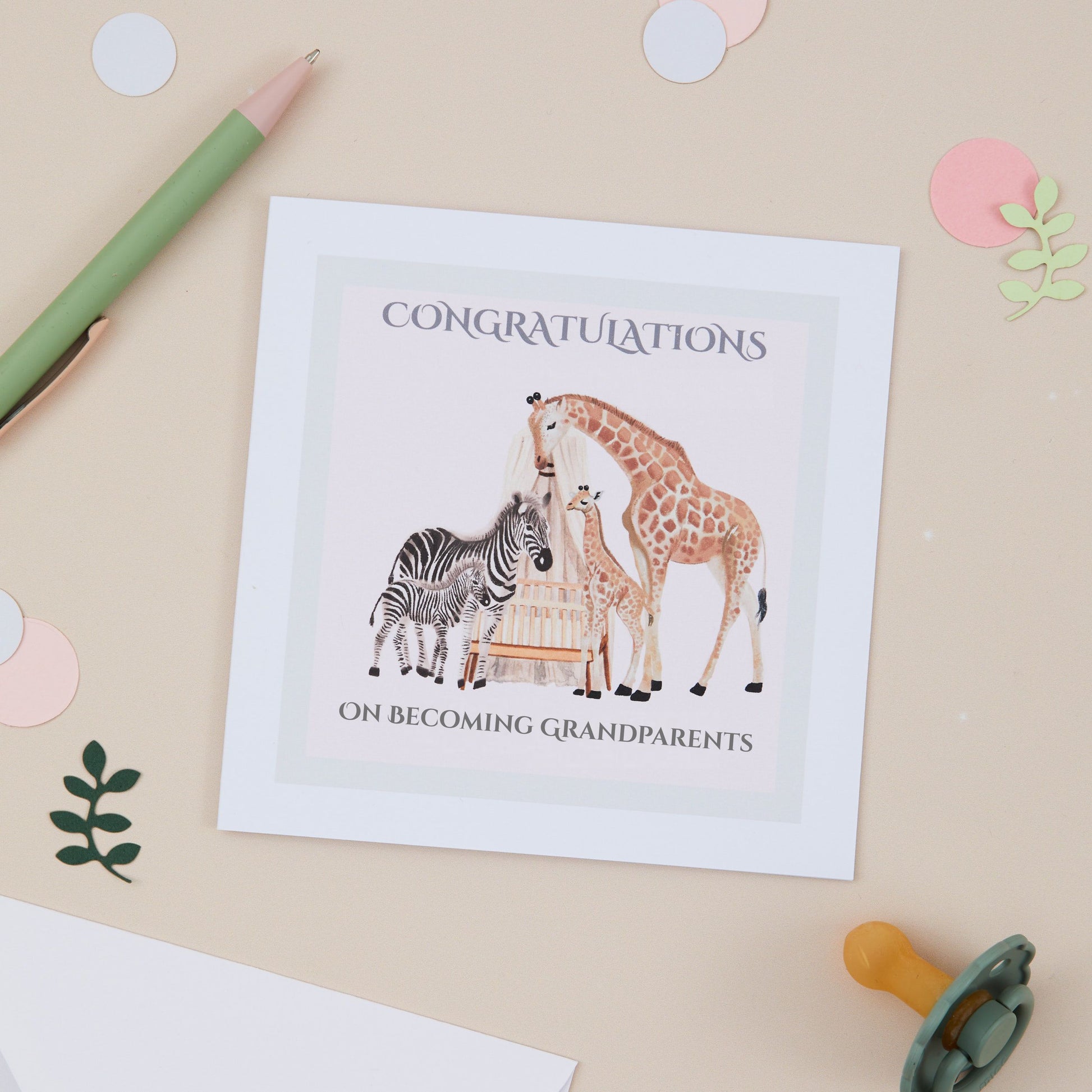 Greeting card with Zebra, Giraffe & their young surrounding a Cot with a pale pink background. The Square card is against a Beige background and there is a pen and a Babies Dummy in the background. The card has the text 'Congratulations on becoming grandparents'.