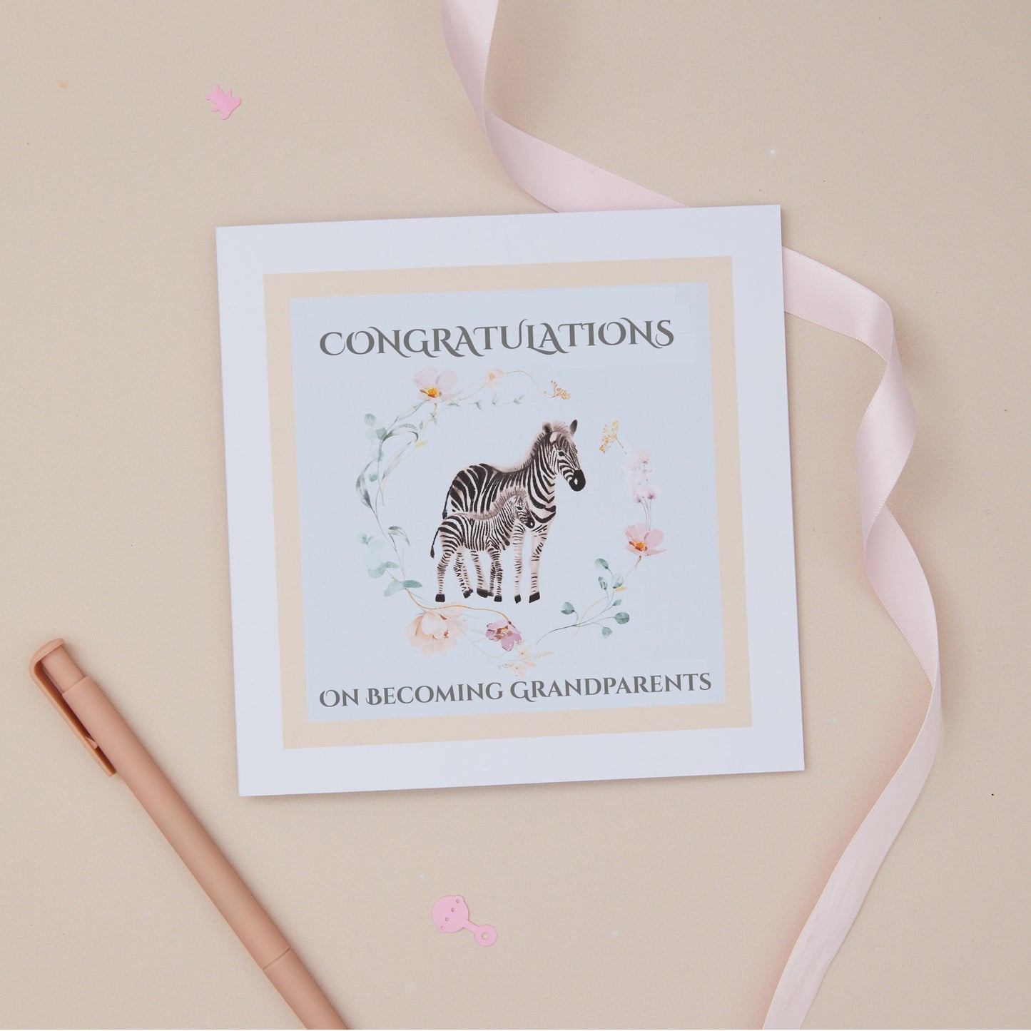 Greeting card with a zebra and its baby illustration within a floral circle on a blue background, and 'Congratulations on becoming grandparents' text. The card is set on a beige background with a ribbon and stationery