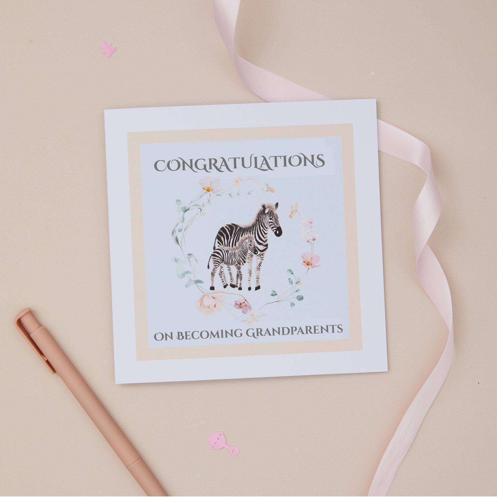 Greeting card with a zebra and its baby illustration within a floral circle on a blue background, and 'Congratulations on becoming grandparents' text. The card is set on a beige background with a ribbon and stationery