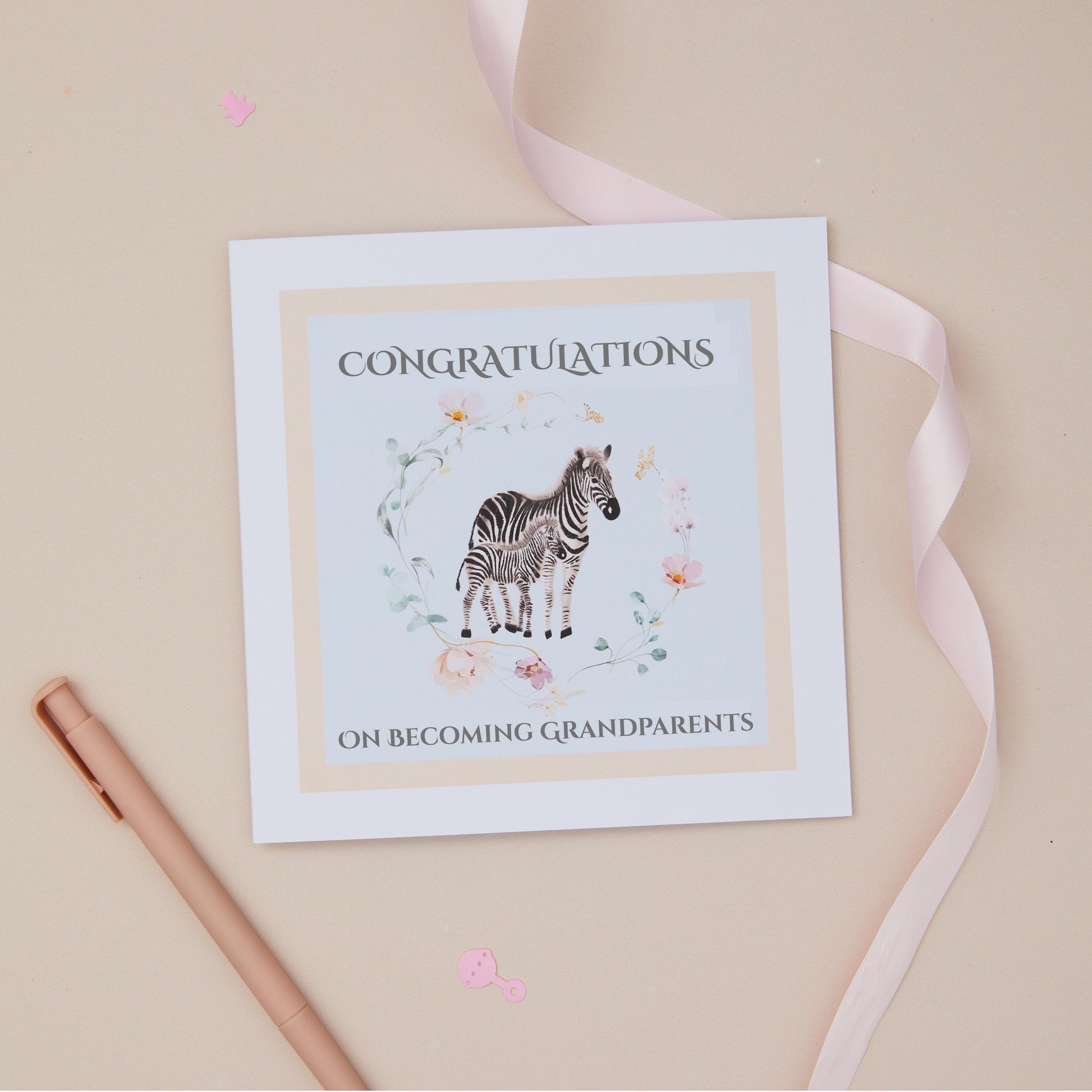 Greeting card with a zebra and its baby illustration within a floral circle on a blue background, and 'Congratulations on becoming grandparents' text. The card is set on a beige background with a ribbon and stationery
