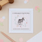 Congratulations on becoming a grandparent Greeting card with a zebra illustration surrounded by a floral circle on a pink background. 'Congratulations on becoming grandparents' text on a beige surface with toys and stationery in the background