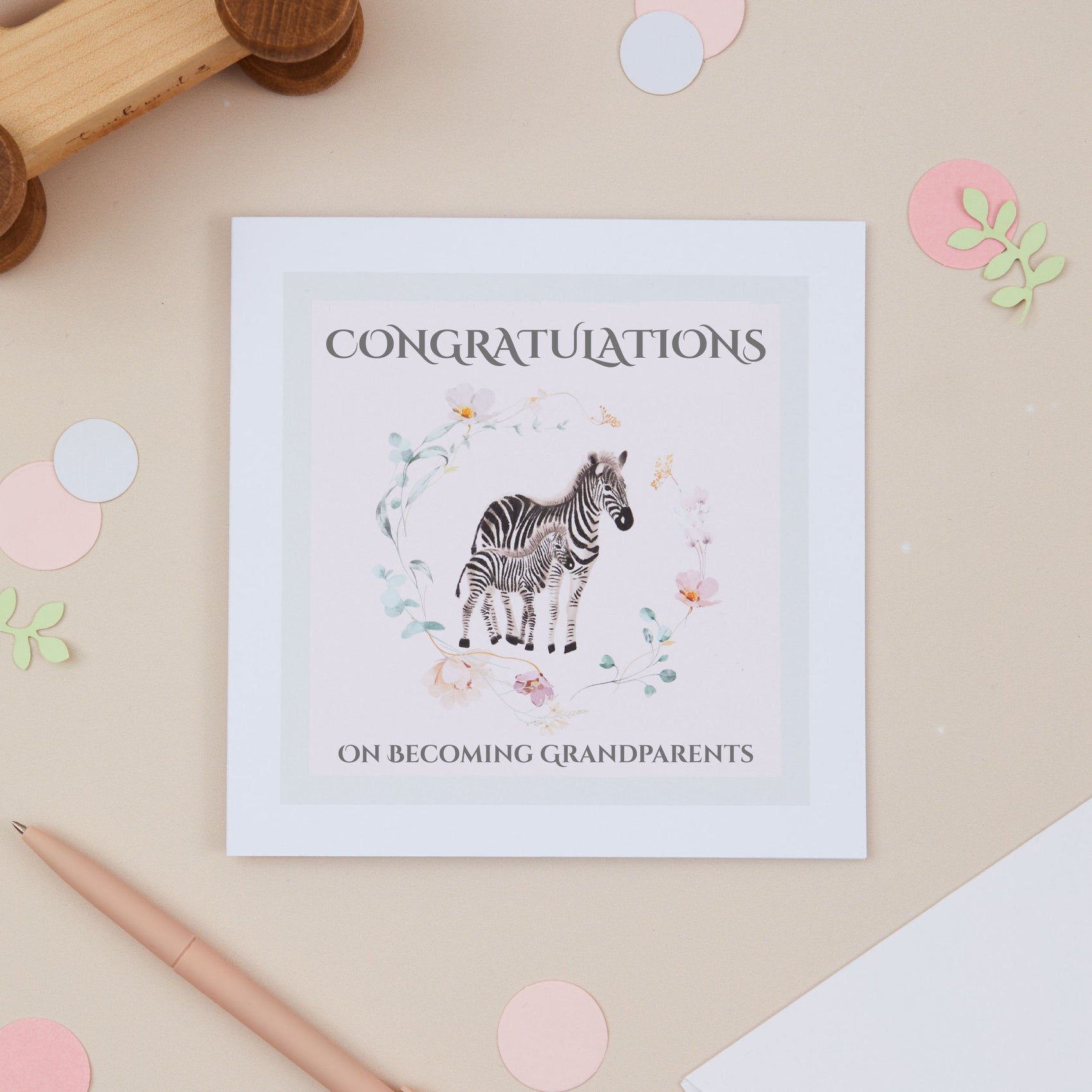 Congratulations on becoming a grandparent Greeting card with a zebra illustration surrounded by a floral circle on a pink background. 'Congratulations on becoming grandparents' text on a beige surface with toys and stationery in the background