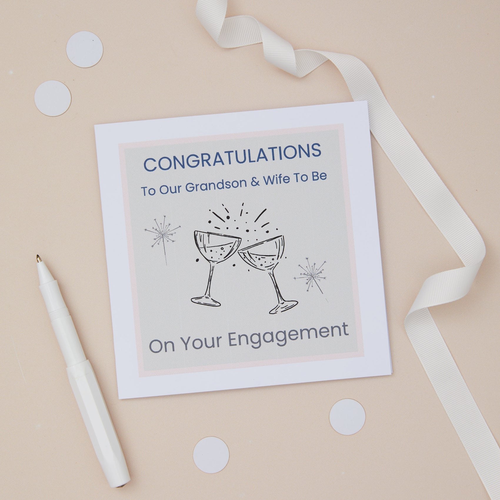 A congratulations card for a grandson's engagement with two celebratory glasses on the front, placed on a neutral background with a white ribbon, pen, and scattered confetti. The text on the card reads "congratulations to our grandson and wife to be on your engagement"