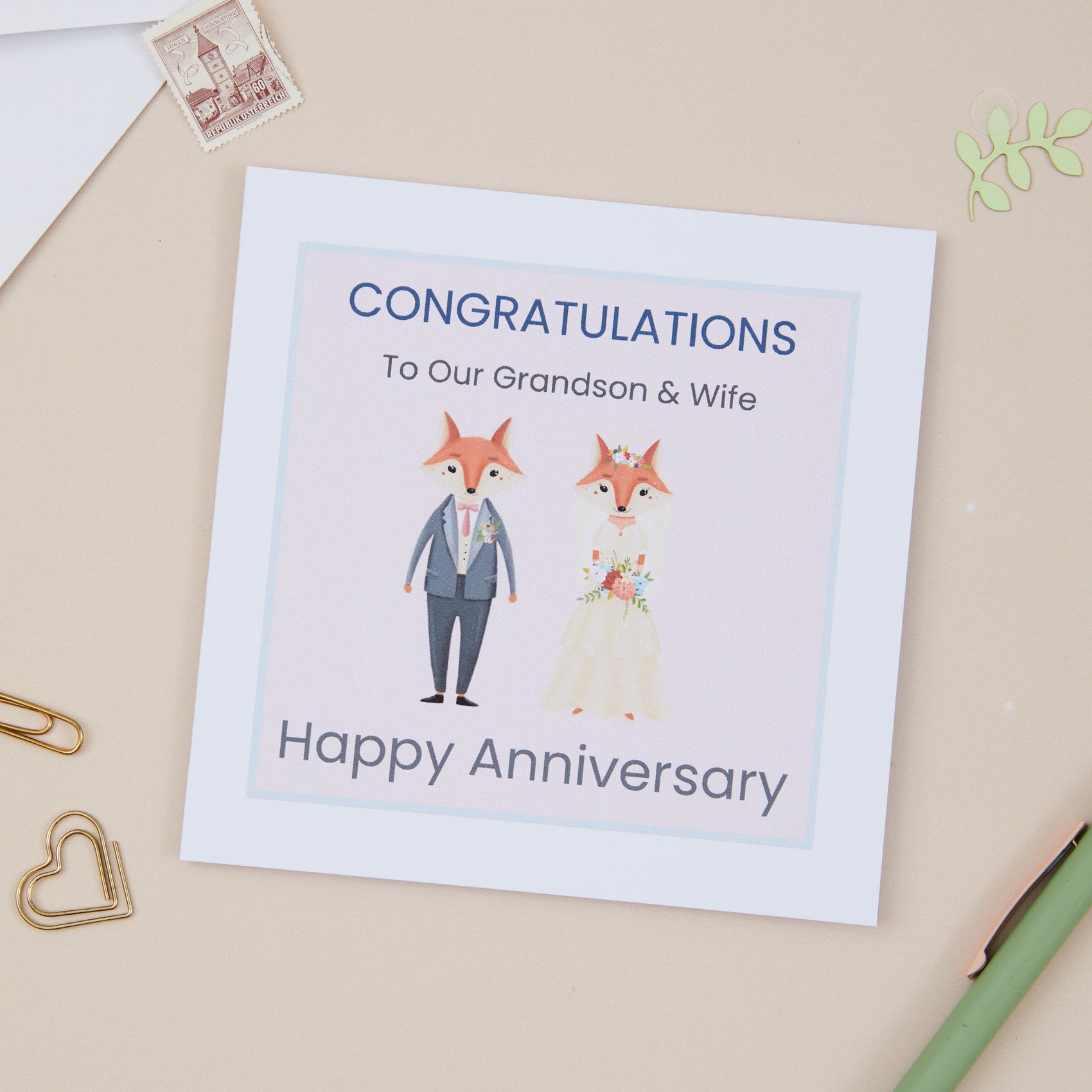 A Grandson Wedding Anniversary Card with an illustration of two foxes dressed in wedding attire, with a 'Congratulations Happy Anniversary' message, placed on a desk with stationery items.