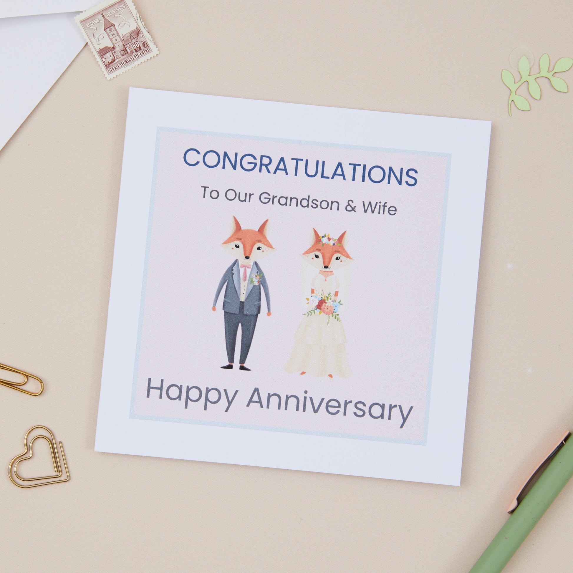 A Grandson Wedding Anniversary Card with an illustration of two foxes dressed in wedding attire, with a 'Congratulations Happy Anniversary' message, placed on a desk with stationery items.