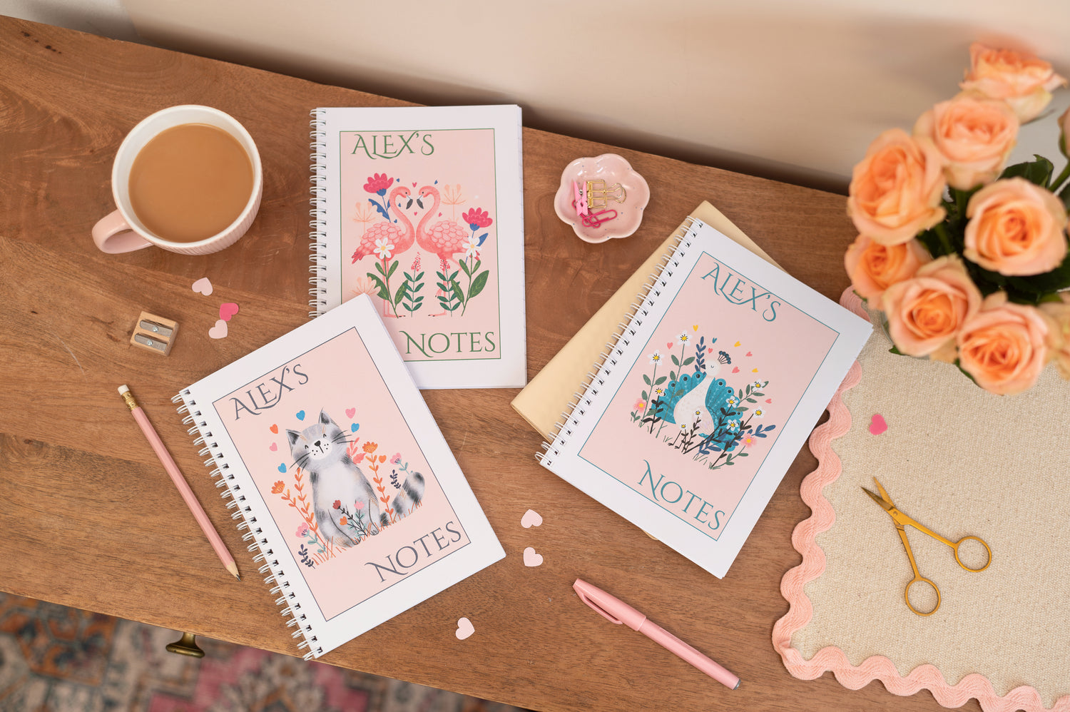 Three notebooks, one has a flamingo pair facing each other, another has a cat, and the other has a peacock. They all have the words that say 'Alex's Notes'. The notebooks are on a table with a cup of tea, flowers and stationery