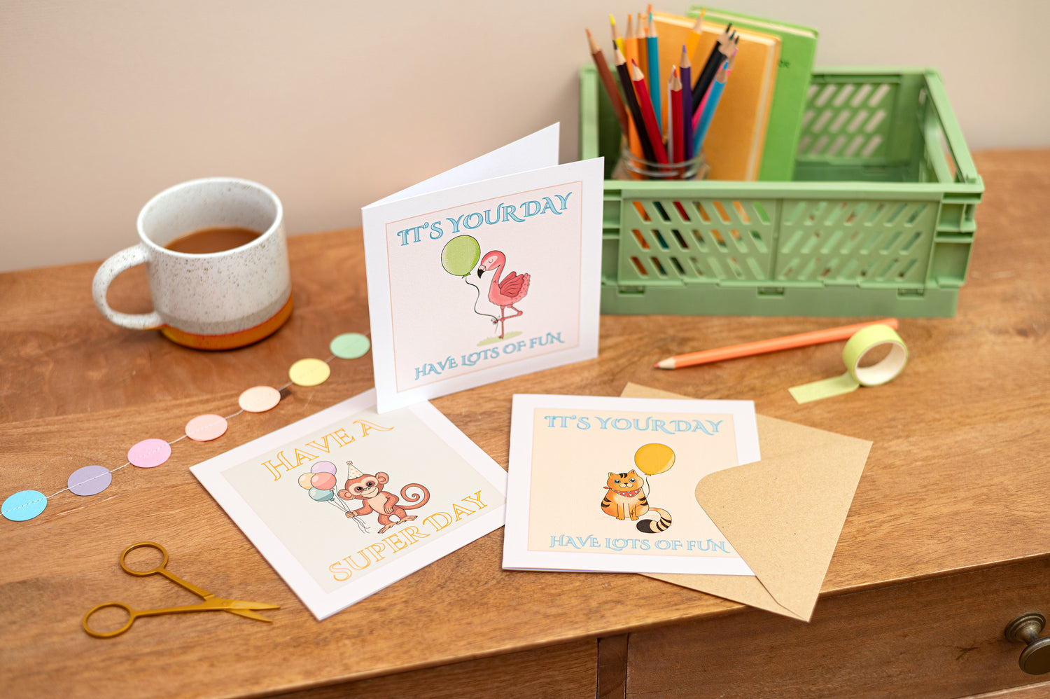 Group Picture of Children's Birthday Cards, featuring the text "it's your day, have lots of fun". The cards sit on a desk with colourful stationery items and a cup of tea. There is also a green box which has some pencils and some post it notes inside it