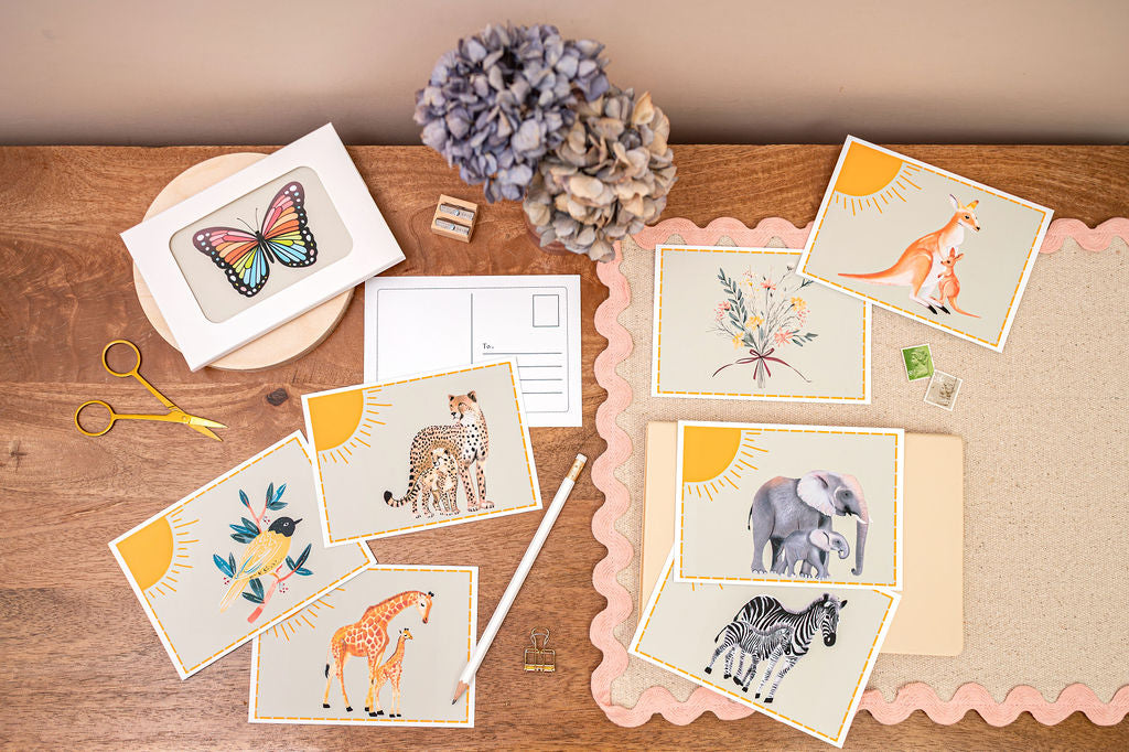 Group photo of A6 Postcard with a Giraffe, Cheetah, Elephant, Kangaroo,  and their Babes, with a yellow sun in the background. The Card is on a desk, surrounded by Stationery.
