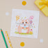 Hamster Kids Birthday Card depicting a colorful illustration of a hamster holding balloons, presented on a table with a yellow envelope, pencils, and scattered confetti.