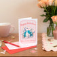 A greeting card with 'Happy Galentine's Day' text and a whimsical peacock illustration , placed on a table with a floral arrangement, a mug, and decorative items.
