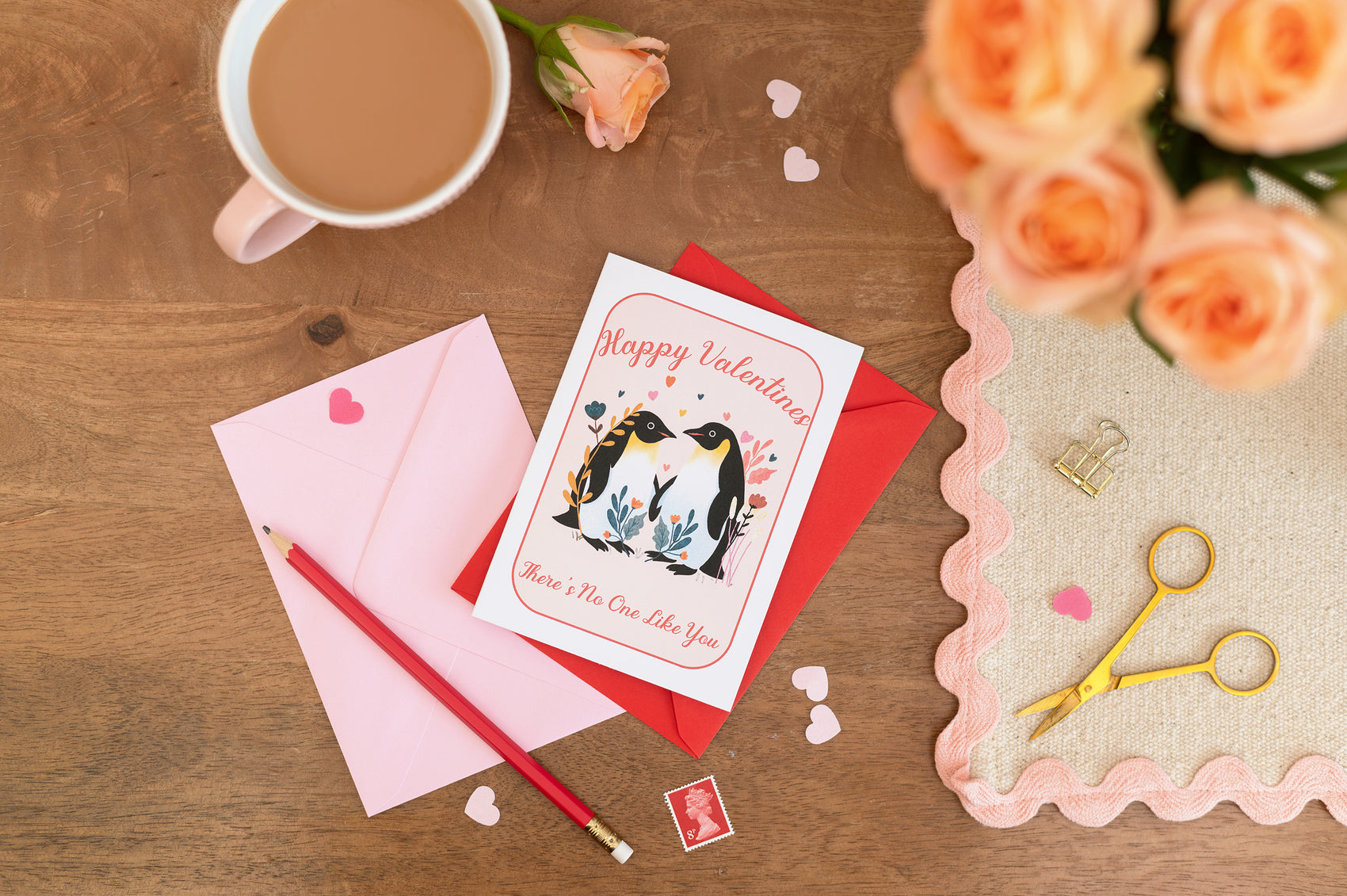 Happy Valentines Penguins Romance Card with pink and red envelopes. The card sits on a desk with stationery and a cup of tea. There is floral roses on the desk