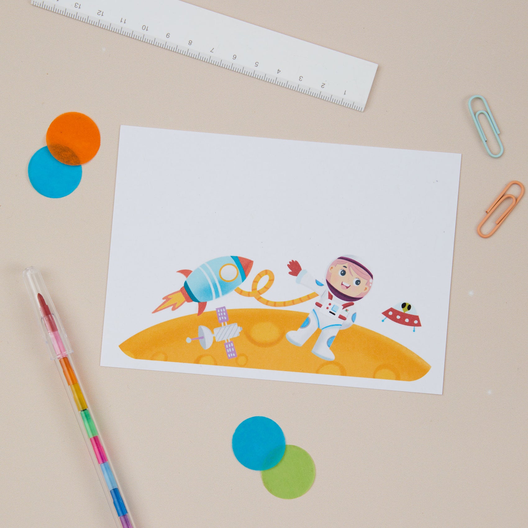 Kids Astronaut Postcard Set. A kids postcard with an astronaut theme, rockets, and planets, accompanied by a pen, ruler, and decorative items on a beige background.