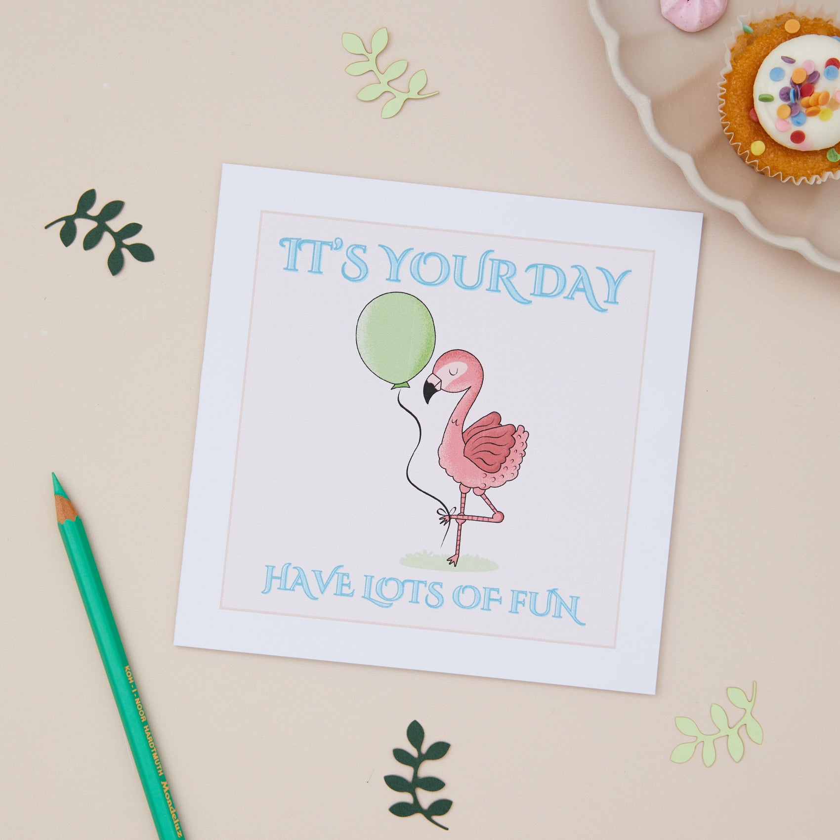 A kids birthday celebration greeting card with a cartoon illustration of a pink flamingo holding a green balloon, with the text 'IT'S YOUR DAY HAVE LOTS OF FUN' on it. The card is displayed on a neutral background with decorative leaves, and a cupcake.