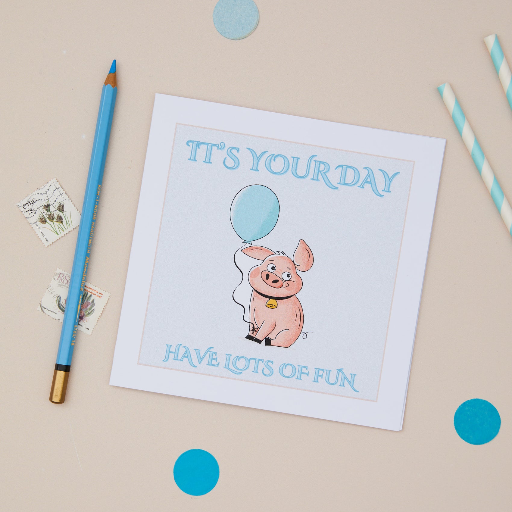 A greeting card with an illustration of a cartoon pig holding a balloon, with the text 'IT'S YOUR DAY HAVE LOTS OF FUN' on it. The card is displayed next to decorative stamps, a pencil, and some decorative blue confetti papers, all placed on a beige background.