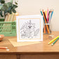 A Kids colouring in greeting card with a raccoon design placed on a table with various coloring pencils surrounding it