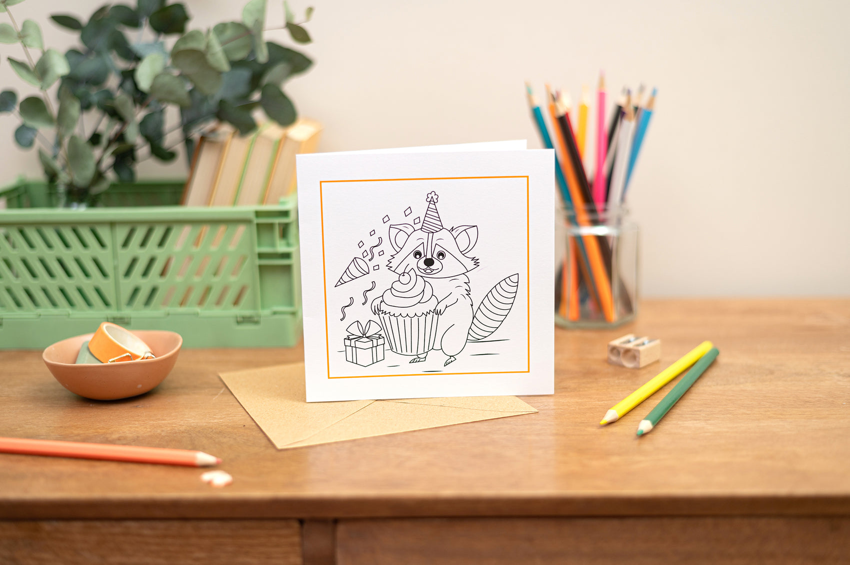 A Kids colouring in greeting card with a raccoon design placed on a table with various coloring pencils surrounding it