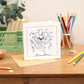 Kids Colouring in Card featuring a party mouse with music notes and party elements around it. The Card is placed on a wooden table with stationery elements surrounding it.