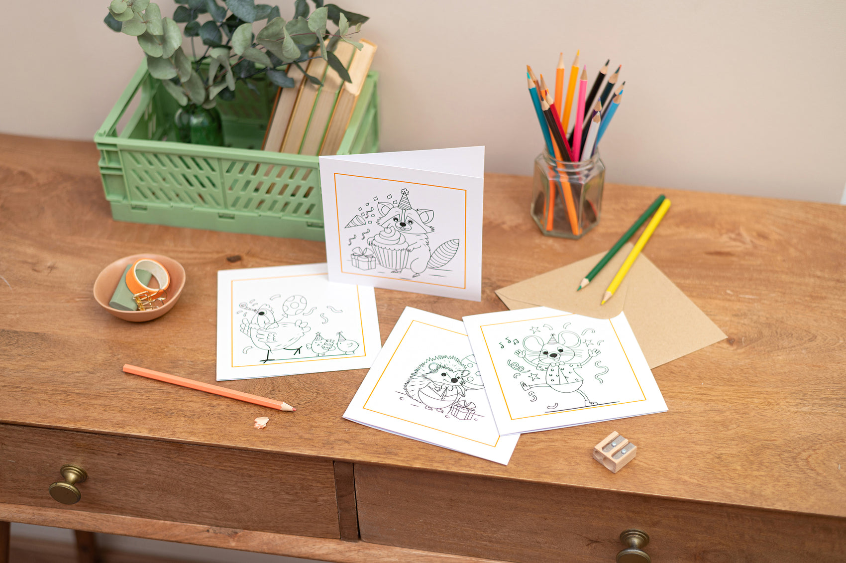 A colleciton of colouring in greeting cards for children, a colouring card of a Raccoon, Party Chicken, Hedgehog and party mouse. The cards sit on a wooden table with stationery pieces surround them