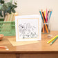 Kids Colouring In Greeting Card featuring a party chicken with chicks in  tow. The front of the card is not coloured so is open to be coloured in. The card sits upright on a wooden table with a green crate, green floral arrangement and stationery.