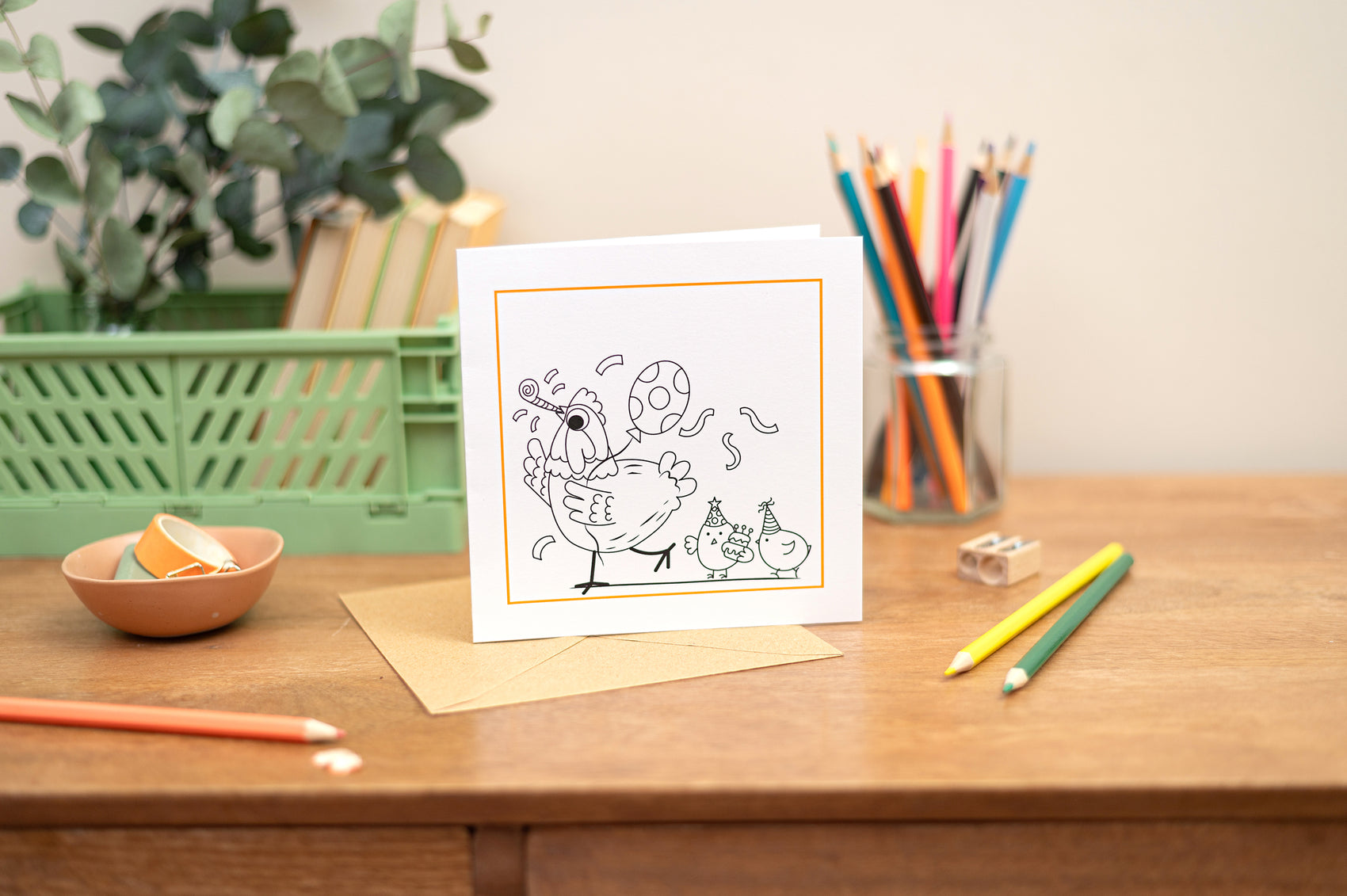 Kids Colouring In Greeting Card featuring a party chicken with chicks in  tow. The front of the card is not coloured so is open to be coloured in. The card sits upright on a wooden table with a green crate, green floral arrangement and stationery.