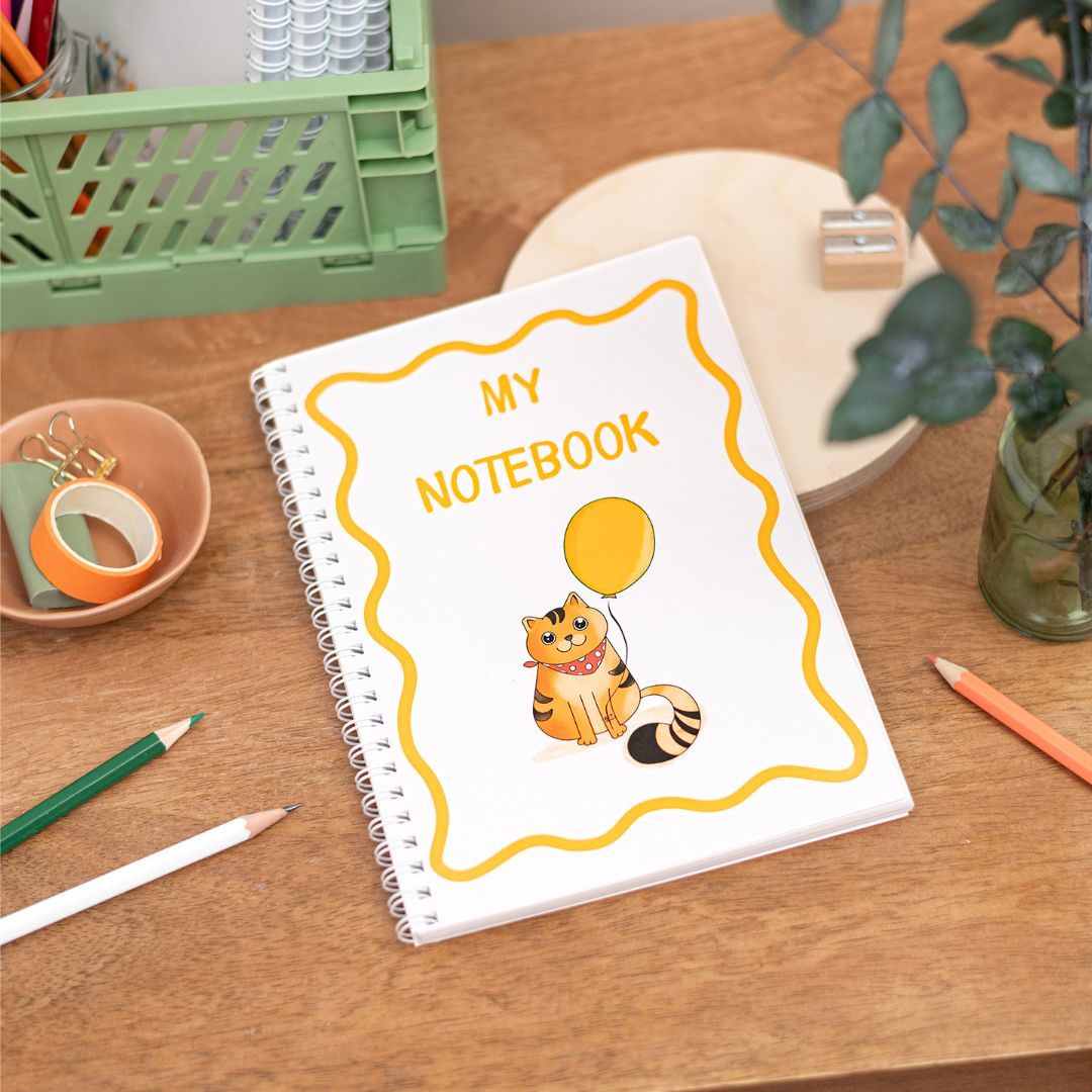 Kids Cute Cat A5 Notebook. A handmade A5 notebook with a white cover featuring a cat illustration, the notebook features the text "my notebook". The notebook is placed on a wooden table alongside stationery items.