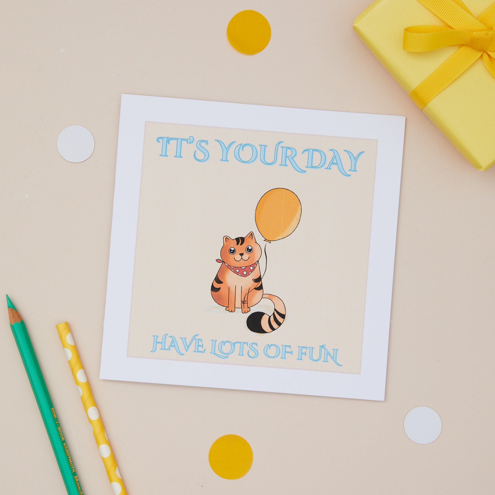 A bright kids birthday greeting card with an illustration of a cat holding a balloon, with the text 'It's Your Day, Have Lots of Fun' displayed. The card is presented against a neutral background with a yellow gift present with a bow, pencils, and decorative dots.