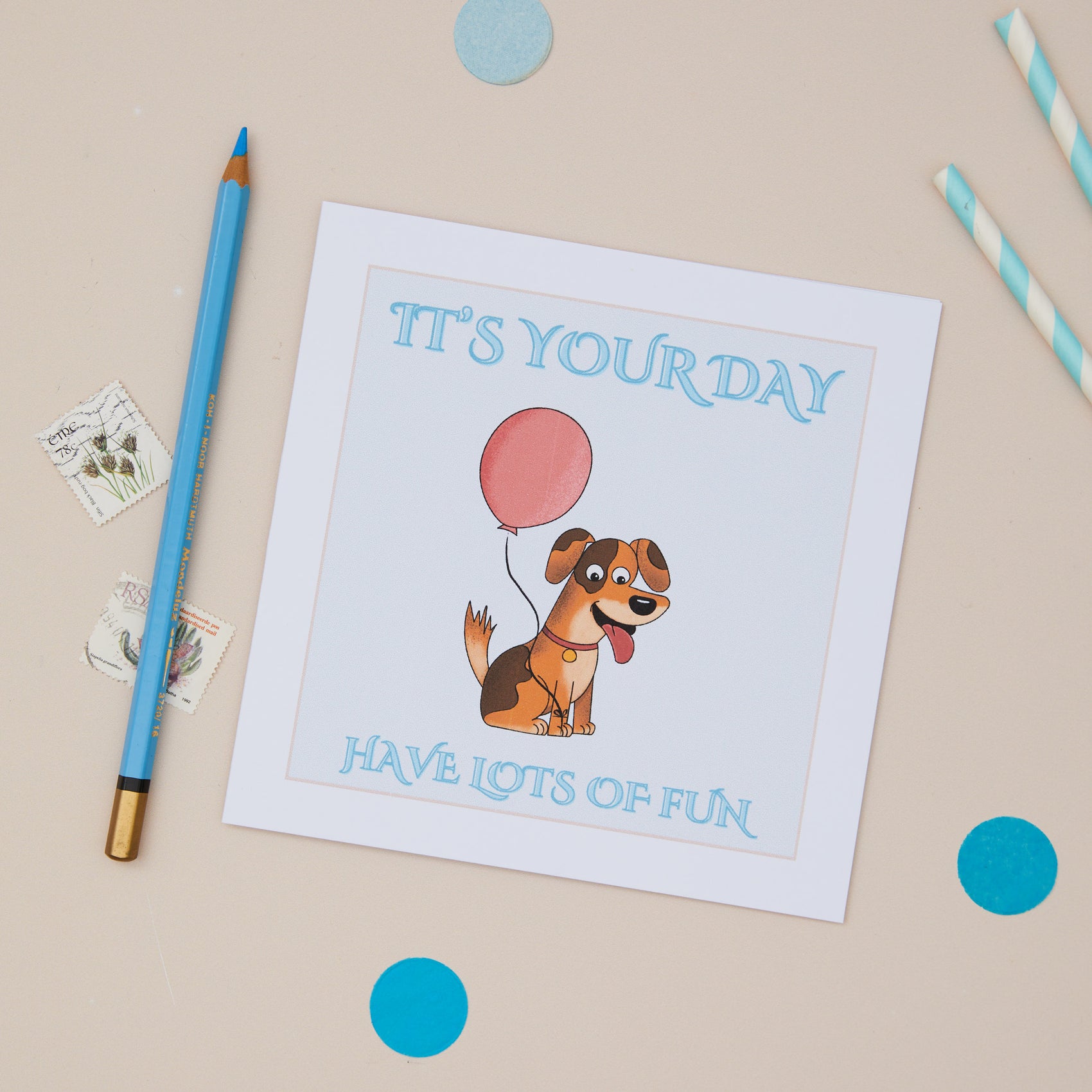 Kids Cute Doggie Birthday Card. The card has an illustration of a dog holding a balloon, with the text 'IT'S YOUR DAY HAVE LOTS OF FUN' on it. The card is placed on a neutral background with a blue pen and decorative elements around it.