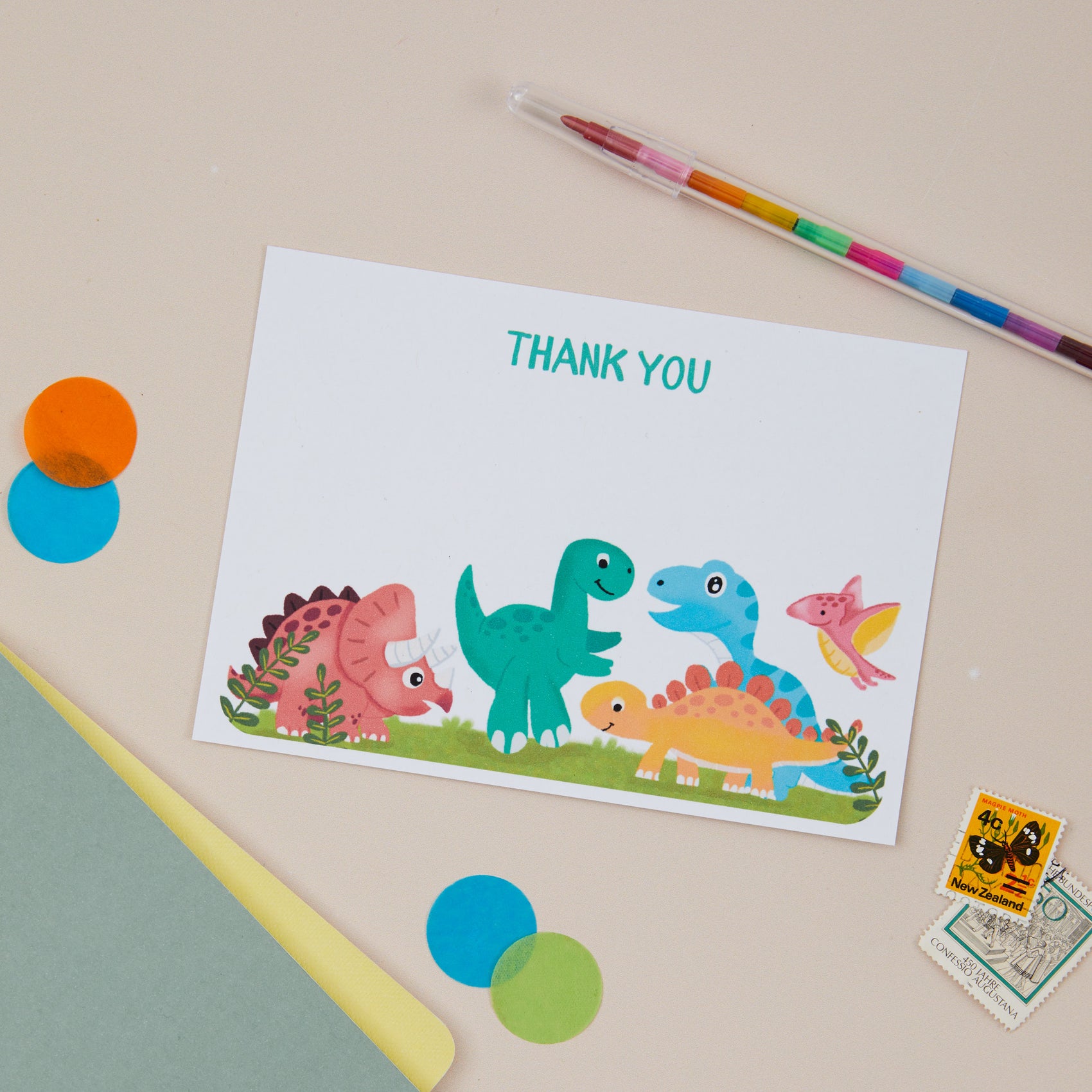 Kids Dinosaur Postcard Set. A flat lay postcard set with illustrations of colourful baby dinosaurs at the bottom. There are decorative elements on a light background like crayons, paper confetti & stamps