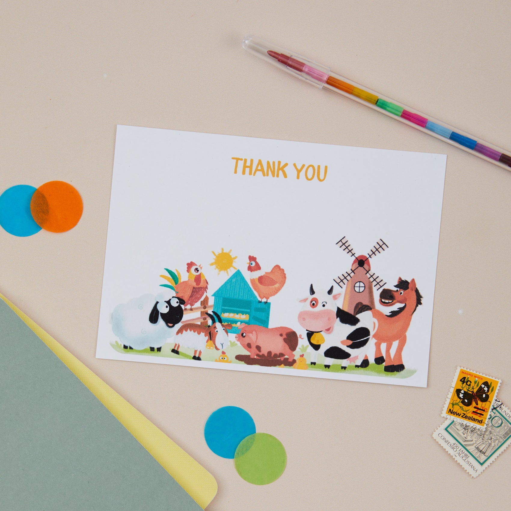 Kids Farmyard Postcard Set. A  postcard with farmyard animal illustrations, including a chicken, pig, and cow, along with a windmill and a blue barn. The card is placed on a desk with colourful circles, a pen, and a stamp.