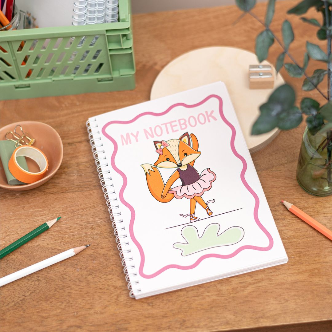 Kids Fox Ballerina A5 Notebook. A handmade A5 notebook with a colourful cover featuring an illustration of a fox ballerina in a tutu. The notebook is displayed on a wooden table alongside pencils and other stationery items.