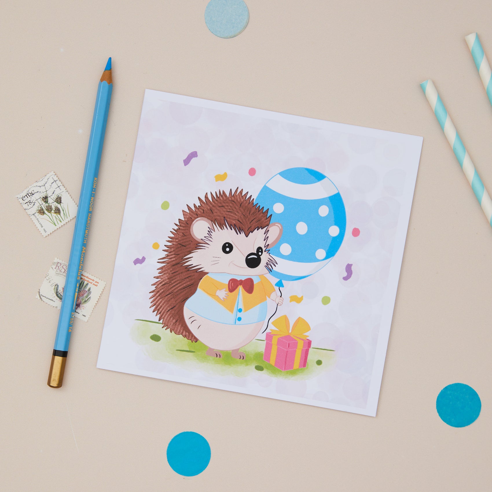 A colourful Party Hedgehog greeting card featuring a cartoon hedgehog with a gift box, and is holding a blue polka dot balloon. The card is displayed on a table with a blue pen and decorative elements.