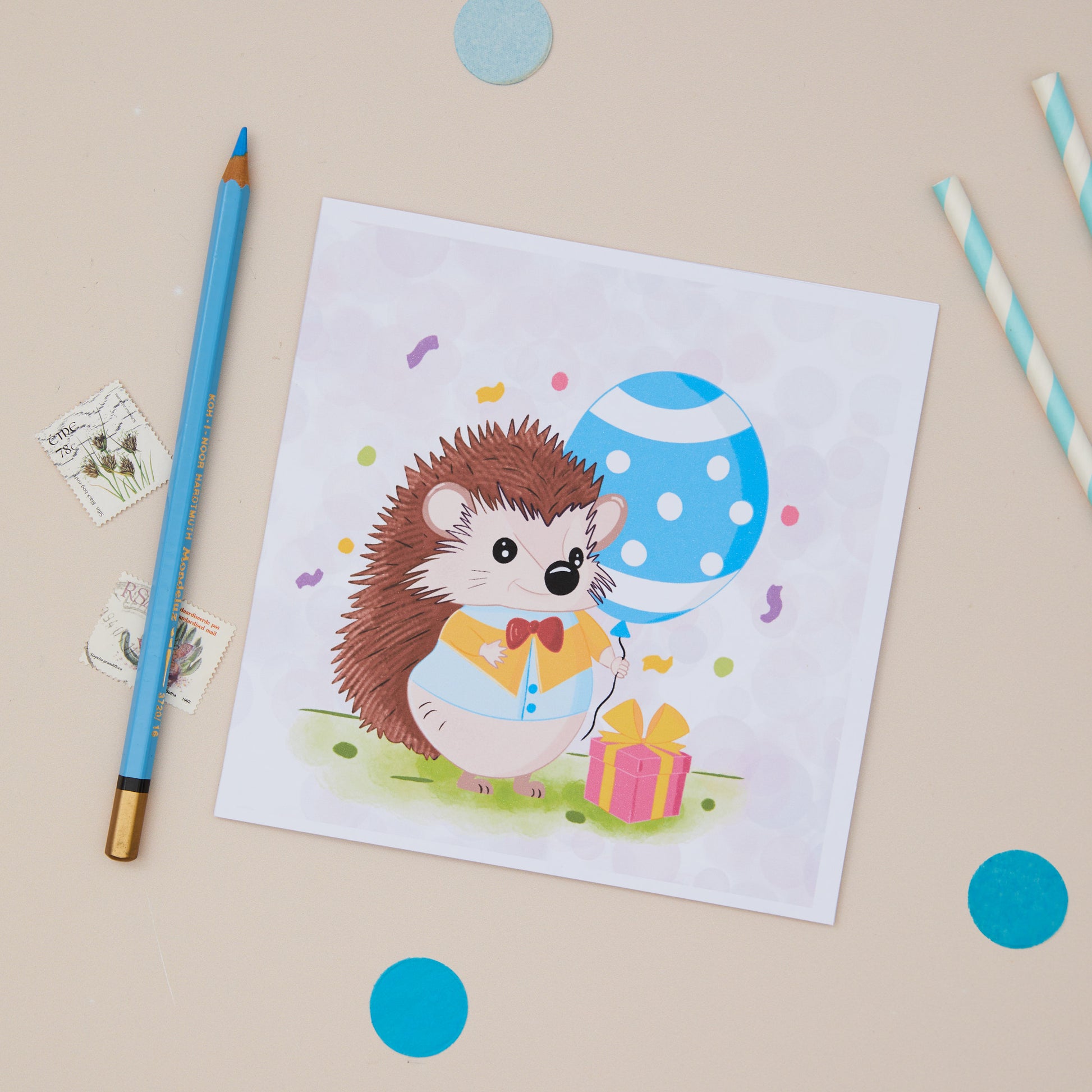 A colourful Party Hedgehog greeting card featuring a cartoon hedgehog with a gift box, and is holding a blue polka dot balloon. The card is displayed on a table with a blue pen and decorative elements.