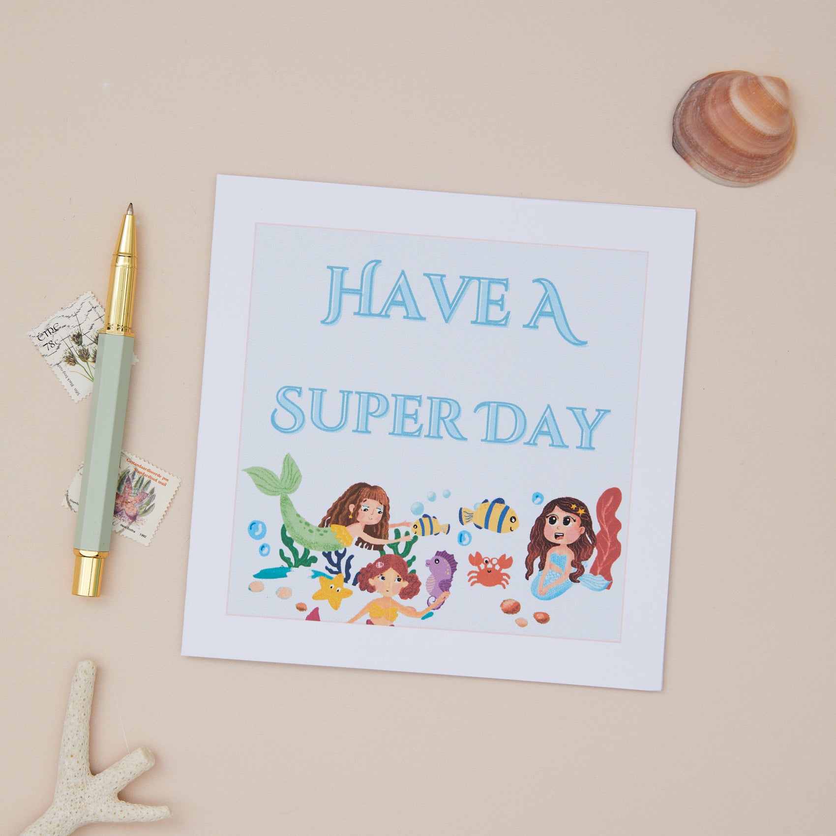 Kids Mermaid Birthday Card with an illustration of mermaids and aquatic life, accompanied by the phrase 'HAVE A SUPER DAY' at the top. The card is displayed on a neutral background with a pen, a shell, and a small decorative butterfly to the side.