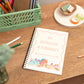 Kids Party Dinosaurs A5 Notebook and Scribbles. A handmade A5 notebook with dinosaur illustrations on the cover, placed on a wooden desk alongside pencils and a vase.