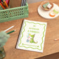 A Kids handmade A5 notebook with a dinosaur illustration on the cover, titled 'MY NOTEBOOK & SCRIBBLES', placed on a wooden desk alongside stationery items.