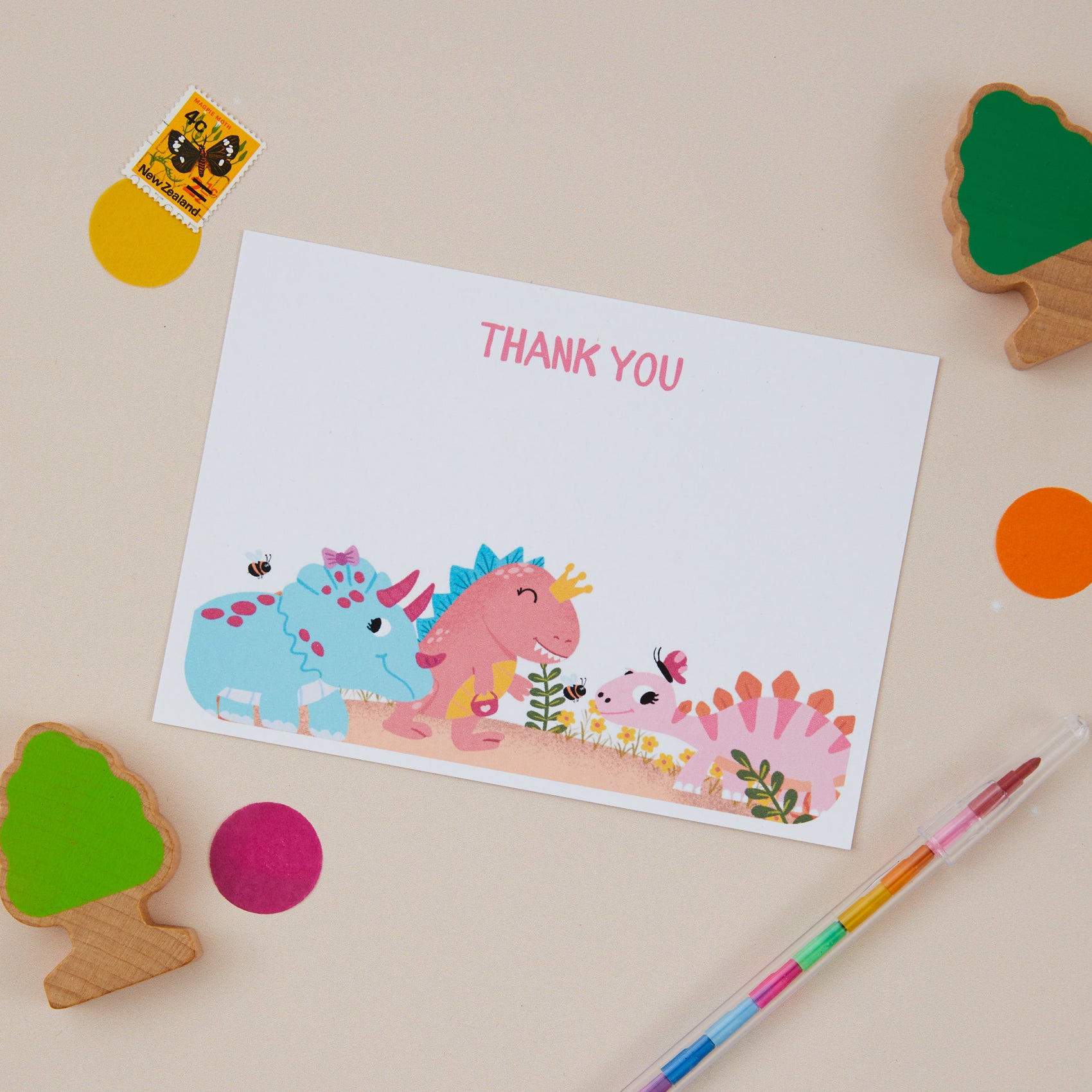 A set of colorful postcards with illustrations of playful dinosaurs at the bottom. The postcards are displayed on a neutral background with craft materials scattered around.
