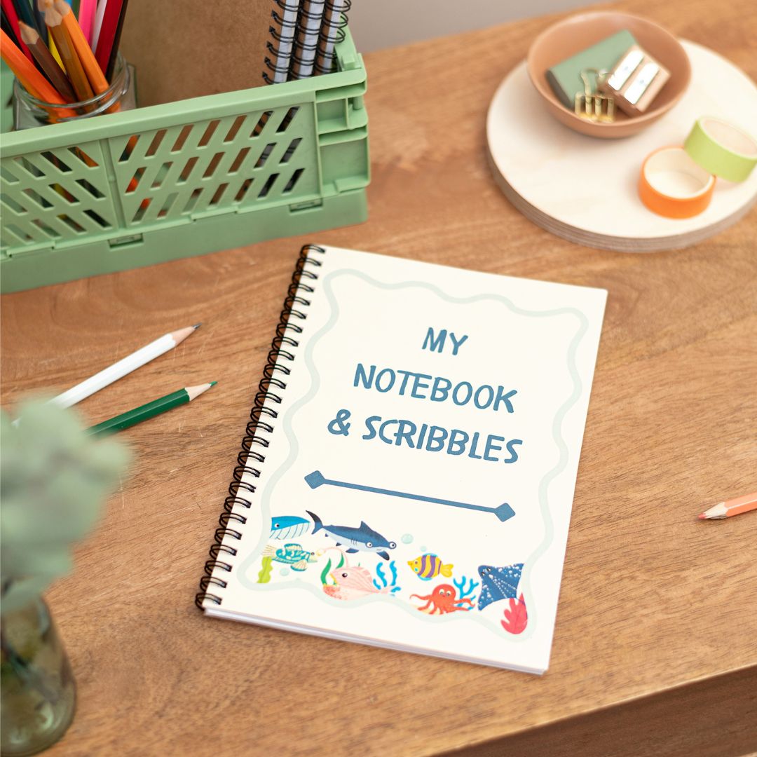 Kids Sealife A5 Notebook. A handmade A5 notebook with a sealife-inspired cover design, titled 'MY NOTEBOOK & SCRIBBLES', placed on a wooden desk alongside stationery items.