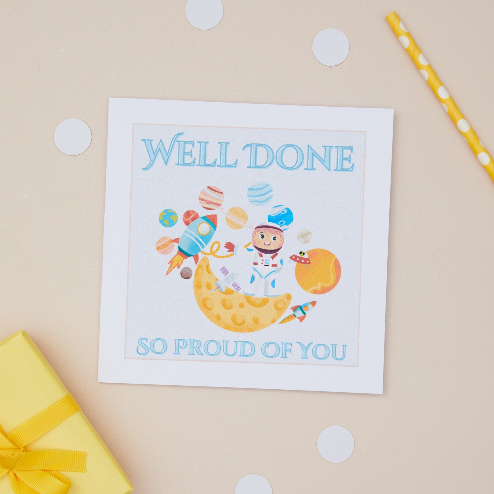 A Kids 'Well Done' card with a cartoon illustration of an astronaut on a star, surrounded by space stars and planets. The Card features the text " well done so proud of you". The card is presented on a background with polka dots.